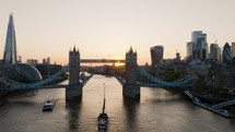 The Sun Is Setting Over The City Of London As A Sail Boat Passes Under Tower Bridge