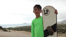 Boy standing with Sand board in tropical island with sand dunes and beach in background - medium shot - serious face