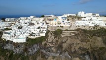 Aerial shot drone medium shot flies toward Thera, Santorini with iconic white buildings on sunny day