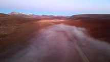 Aerial shot drone flies over red desert with snow capped mountains away from fog covered valley just before sunrise