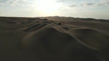Aerial shot drone flies toward sun over sand dunes and silhouettes of dune buggy and hikers near desert oasis Huacachina, Peru