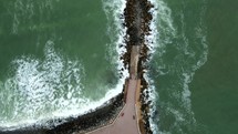 Aerial shot drone flies forward with camera facing down over rocky pier jutting into sea with waves crashing on it