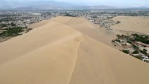 Aerial shot drone flies over ridge of sand dune above and to left of desert oasis in Huacachina, Peru