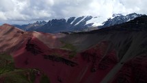 Aerial shot drone hovers in front of red and gray mountain with snow capped mountains in the background