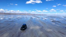 Aerial shot drone flies backwards in front of jeep driving on salt flats with reflective water showing a blue sky with fluffy white clouds