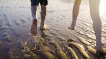 Children's Feet Running Through Water On The Beach Together In Warm Summer Sun Light