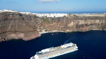 Aerial shot drone flies over cruise ship docked in Santorini toward cable car at base of iconic city with white buildings and blue domes