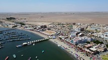 Aerial shot drone orbits to the right over boats in the harbor and sumbrellas on the beach in the desert town of Paracas, Peru