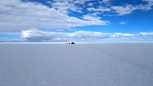 Aerial shot drone flies low to ground and orbits to left around jeep in middle of white salt flats contrasted by blue sky