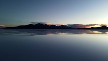 Aerial shot drone descends over salt flats covered in water with reflections of mountain range in front of sunset