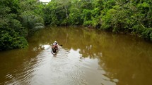 Aerial shot drone flying low over water following canoe full of people paddling down brown river in middle of Amazon rainforest