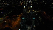 Aerial shot drone flies forward from above and behind the Basilica del Voto Nacional with a nighttime city of Quito in background
