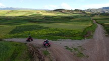 Aerial shot drone follows five quad bikes driving through green fields on a dirt road