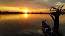 Aerial shot drone flies forward toward silhouetted tree and canoe boat on black lagoon in middle of Amazon rainforest at sunset