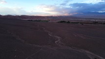 Aerial shot drone flies forward away from sunset over red desert toward green oasis