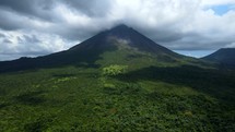 Aerial shot drone flies forward toward volcano in middle of lush green forest with clouds casting shadows on landscape
