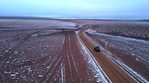 Aerial shot drone follows jeep through red desert with fresh snow on ground just before sunrise