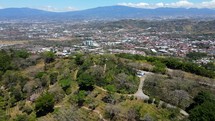 Aerial shot drone flies over hill at top of coffee plantation toward city with mountains behind it
