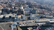 Aerial shot drone flies forward with camera angled down over loading docks and main plaza next docks in coastal city on sunny day
