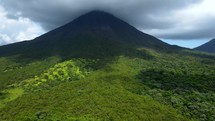 Aerial shot drone slowly lowers in front of volcano under cloud cover surrounded by lush green green jungle in sunlight