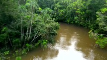 Aerial shot drone rising up as canoe full of people paddling down brown river in middle of Amazon rainforest are revealed from behind trees