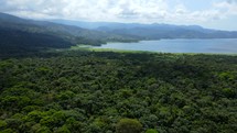 Aerial shot drone flies forward over lush green forest toward blue lake near volcano