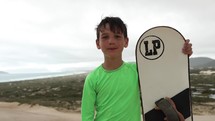 Young boy holding up sand board in front of sand dunes and beach in background - medium shot