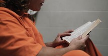 A sad man, convicted prisoner inmate wearing an orange prison uniform/jumpsuit sitting in his prison cell reading a bible.