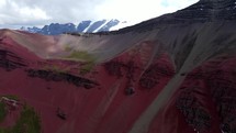 Aerial shot drone ascends over red and gray mountain with snow capped mountains in the background