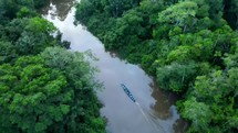 Aerial shot drone slowly following motorized canoe boat going down brown river in middle of Amazon rainforest