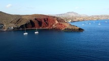 Aerial shot drone flies to the right at Red Beach with two sailboats in bay in the south of Santorini
