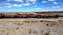 Aerial shot drone flies up and away from train cemetery in middle of desert with adjacent town in the background
