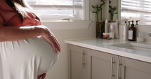 9 month pregnant woman looks over herself and belly in bathroom mirror - side profile