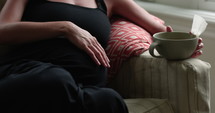 Pregnant woman sits on couch sipping on tea 