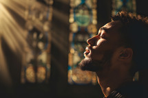 young Man worshiping in prayer in a church, eyes closed, sunshine coming through window onto his skin, space for text