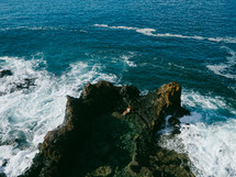 A woman in a swimsuit sits on craggy rocks surrounded by ocean waves.