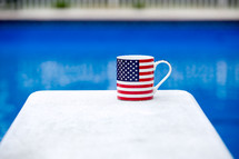 A flag coffee mug on a white table next to a blue swimming pool.