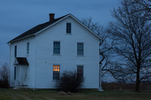 White farm house at dusk with one lit window