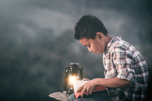 A boy child reading a Bible by a lantern outdoors 