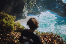 man looking down at a waterfall off a cliff on a beach near the ocean 