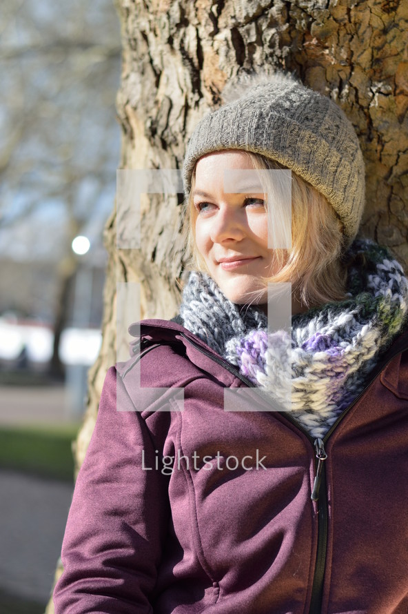 a woman in a beanie leaning against a tree 