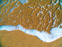 Sea foam from an ocean wave  sweeps across the sand at a beach with grains of sand and pebbles.  The Earth's Oceans are a great resource of life for this planet and need to be protected now more than ever. 