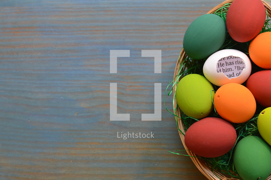 He is Risen scripture in brightly colored Easter Eggs in a basket on cyan wood background 