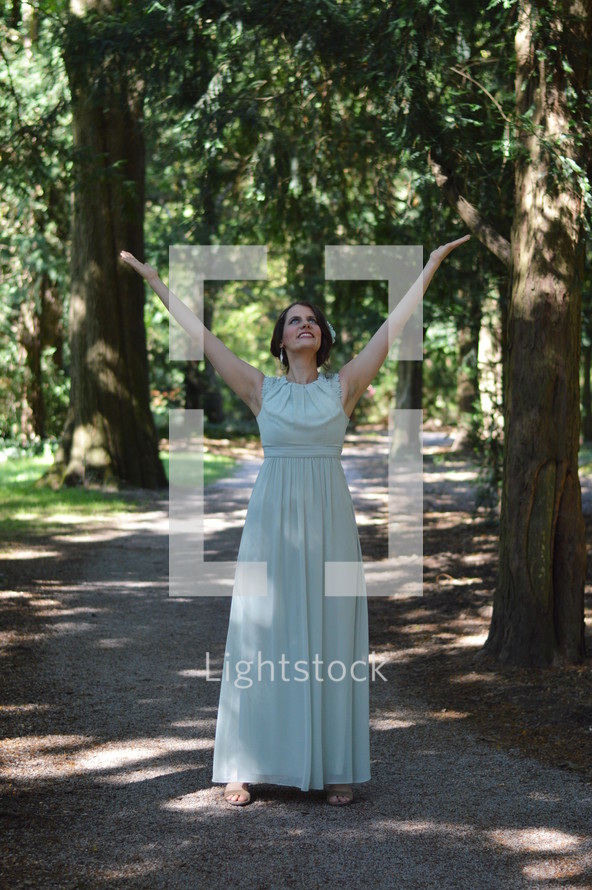 a woman in a long dress standing on a path in a forest with raised hands 