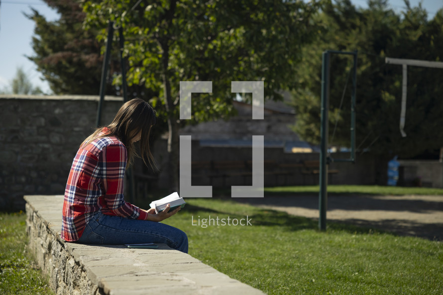 Young woman sitting outside on rock wall reading her Bible