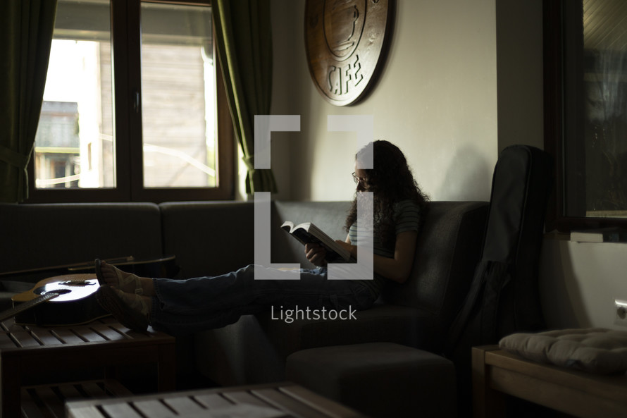 Young woman sitting in a dark room in front of window reading her Bible