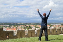woman with raised arms in adoration standing on top of the walls of an old fortress looking over a town
