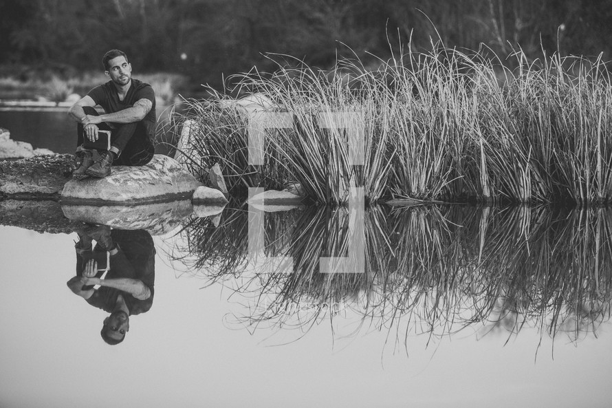 A man sitting at the shore of a pond with a Bible 