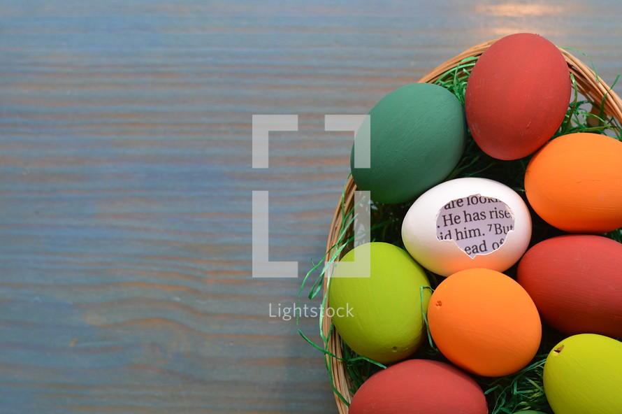 He is Risen scripture in brightly colored Easter Eggs in an Easter basket on cyan wood background 