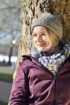 a woman in a beanie leaning against a tree 